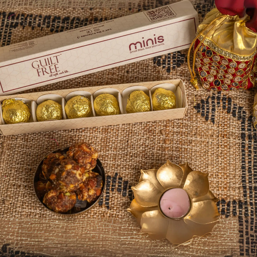 Box of gold-wrapped with 'minis' branding, laddus, and a decorative candle on a textured surface.