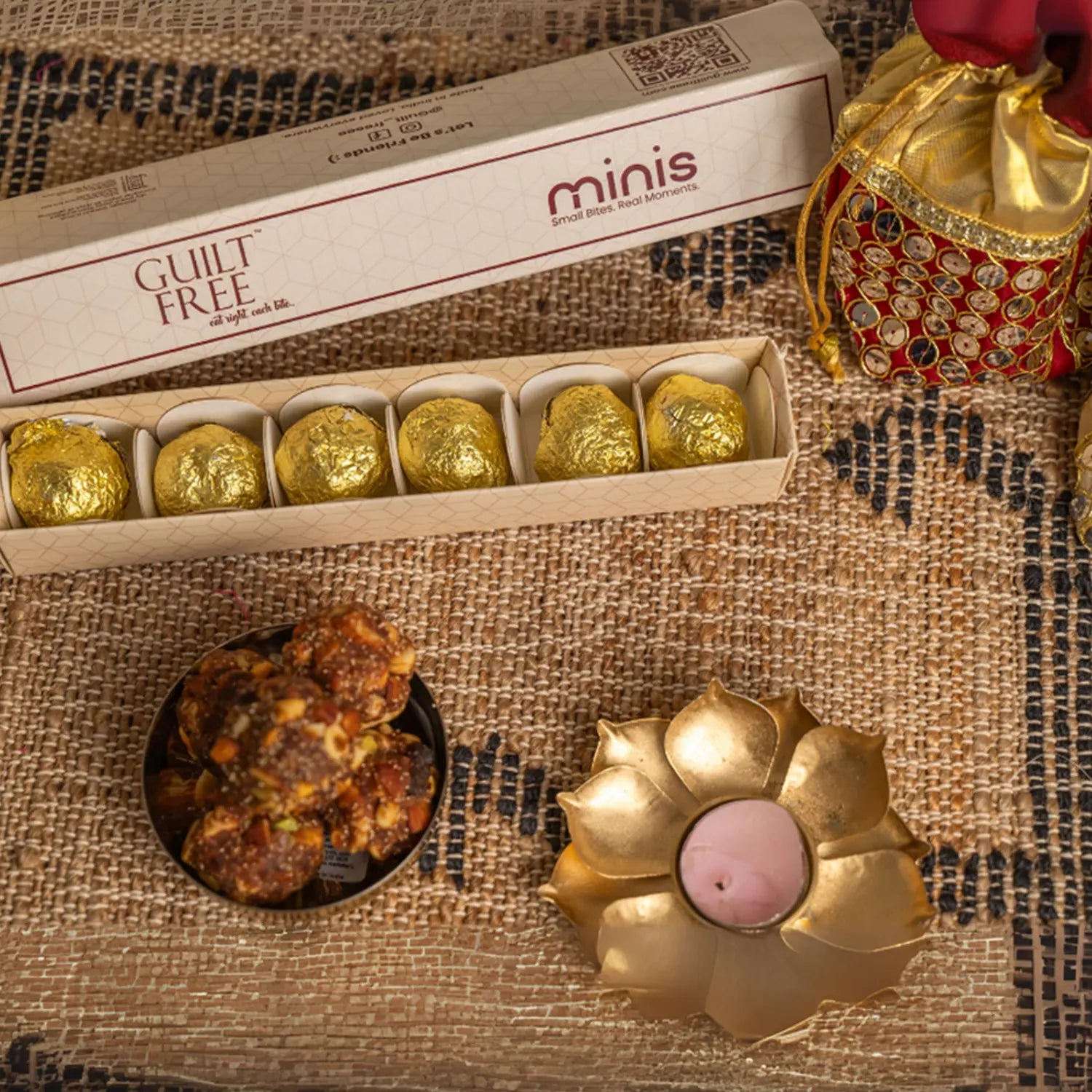 Box of gold-wrapped with 'minis' branding, laddus, and a decorative candle on a textured surface.